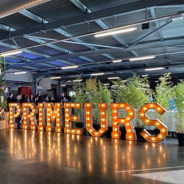 Große, beleuchtete Buchstaben spelling -PRIMEURS- stehen in einem modernen Raum, umgeben von kleinen Pflanzen. Im Hintergrund sind Menschen und Stände sichtbar, die auf eine Veranstaltung hinweisen.