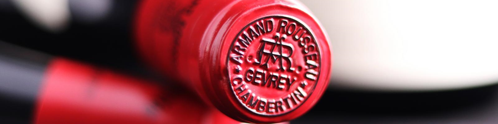 Domaine Armand Rousseau Ein runder, roter Korken mit einer eingeprägten Siegelmarke. Der Korken stammt von einer Weinflasche, im Hintergrund unscharfe Flaschen und eine helle Umgebung.