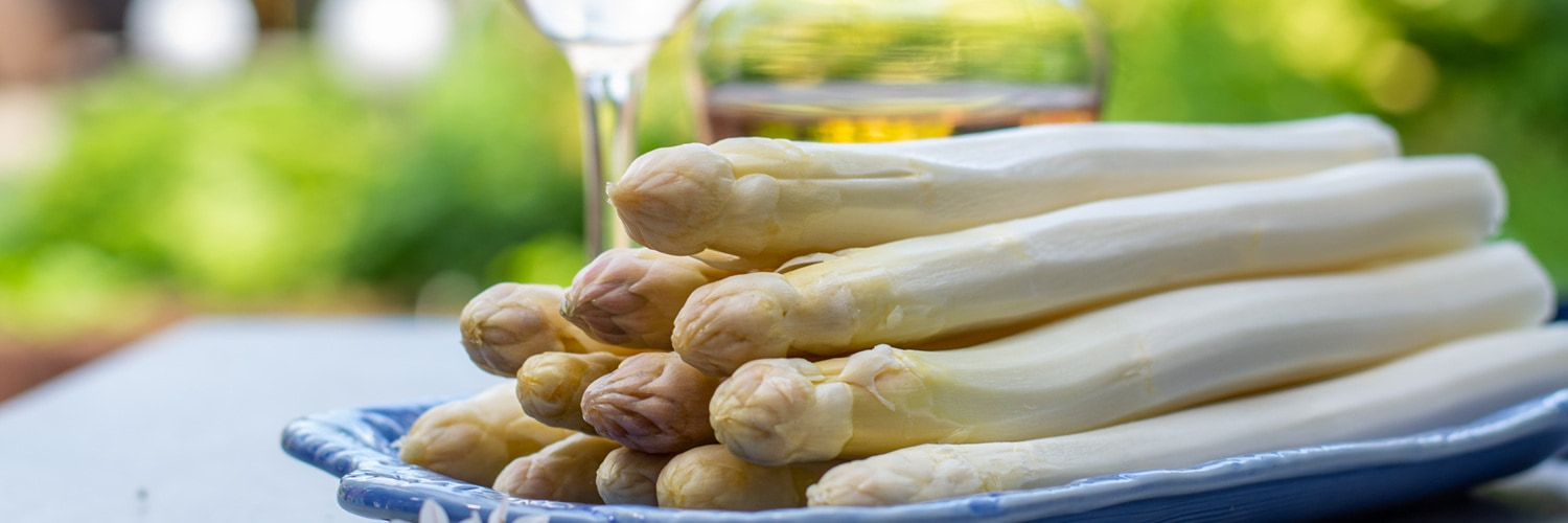 Eine schwarze Pfanne liegt auf einem Holztisch. Daneben steht eine Flasche Wein und ein Glas. Ein gebündelter Spargel mit einer Schleife aus Jute ist ebenfalls sichtbar.