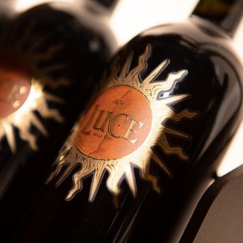 Schwarze Weinflaschen stehen aufrecht in einem Halter. Sie haben ein auffälliges, runde Etikett mit goldenen und orangen Sonnenstrahlen, das den Namen -LUCE- zeigt.