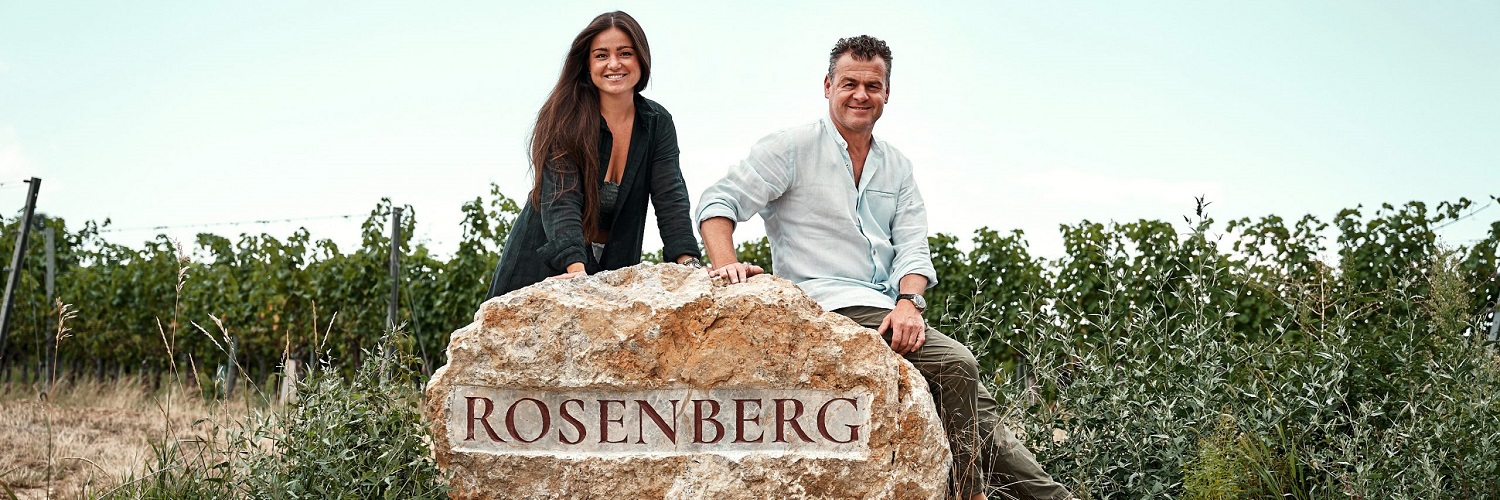 Auf einen Wein mit... Johanna Markowitsch Zwei Personen sitzen auf einem großen Stein mit der Aufschrift -ROSENBERG-. Im Hintergrund sind Weinreben und ein klarer Himmel sichtbar. Die Szene vermittelt eine ländliche, entspannte Atmosphäre.