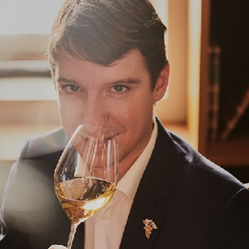 Ein Mann in einem dunklen Anzug hält ein Weinglas vor sein Gesicht und lächelt geheimnisvoll. Der Hintergrund zeigt eine gemütliche, gut beleuchtete Umgebung mit Bücherregalen.