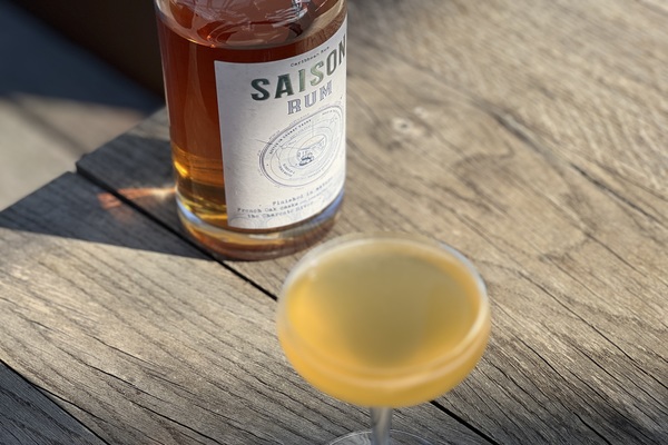 Ein Glas mit einem gelben Cocktail steht auf einem Holztisch, während eine Flasche Saison Rum im Hintergrund sichtbar ist. Der Tisch ist hell beleuchtet.