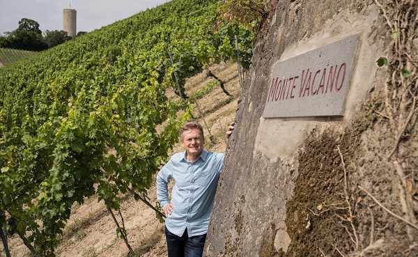 Ein Mann steht in einem Weinberg, posierend neben einem Stein mit der Aufschrift -Monte Vacano-. Im Hintergrund sind grüne Weinreben und ein Aussichtsturm zu sehen, der sich auf einem Hügel befindet.