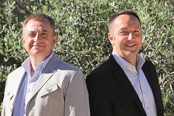 Zwei Männer stehen Rücken an Rücken, lächeln und tragen formelle Kleidung. Sie sind vor grünen Olivenbaumblättern positioniert, was eine sonnige, natürliche Umgebung schafft.