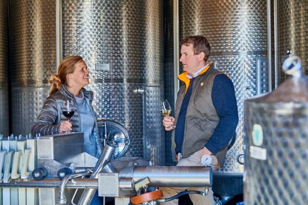 Ein Paar steht in einem Weinkeller, lächelt sich an und hält Weingläser. Der Hintergrund besteht aus großen, glänzenden Edelstahltanks, während eine Weinpressmaschine im Vordergrund sichtbar ist.