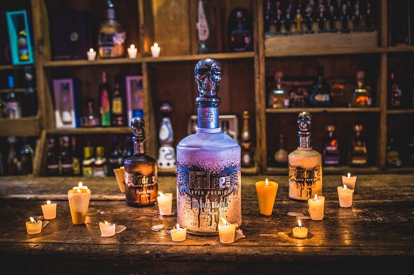 Auf einem Holztisch stehen drei bedeutende Flaschen Tequila, umgeben von kleinen Kerzen. Im Hintergrund sind Regale mit weiteren Spirituosen sichtbar, während sanftes Licht eine gemütliche Atmosphäre schafft.
