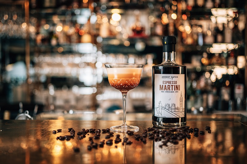 Ein Glas Espresso Martini steht auf einer Bar, umgeben von Kaffeebohnen. Im Hintergrund sind verschwommen Flaschen und Gläser in einem stilvollen Barambiente sichtbar.