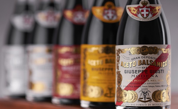 Sechs Flaschen Balsamico-Essig stehen in einer Reihe, jede mit verschiedenen Etiketten und Farben. Die Flasche im Vordergrund hat ein goldnes und rotes Design mit dem Text -ACETO BALSAMICO DI GIUSEPPE GIUSTI MODENA-.