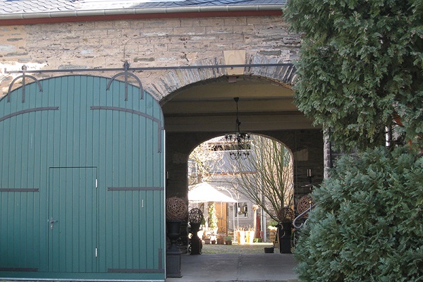 Ein großer, grüner Torbogen öffnet sich zu einem inneren Hof mit verschiedenen Pflanzen und Möbeln. Auf der linken Seite sind große Bäume und eine steinerne Wand zu sehen.