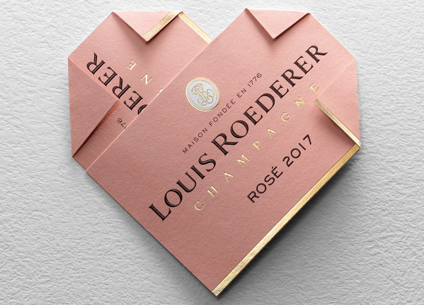 Zwei ineinandergreifende rosafarbene Weinetiketten bilden ein Herz. Die Etiketten tragen den Schriftzug -LOUIS ROEDERER CHAMPAGNE ROSÉ 2017- in eleganter Schriftart, ergänzt durch goldene Akzente.