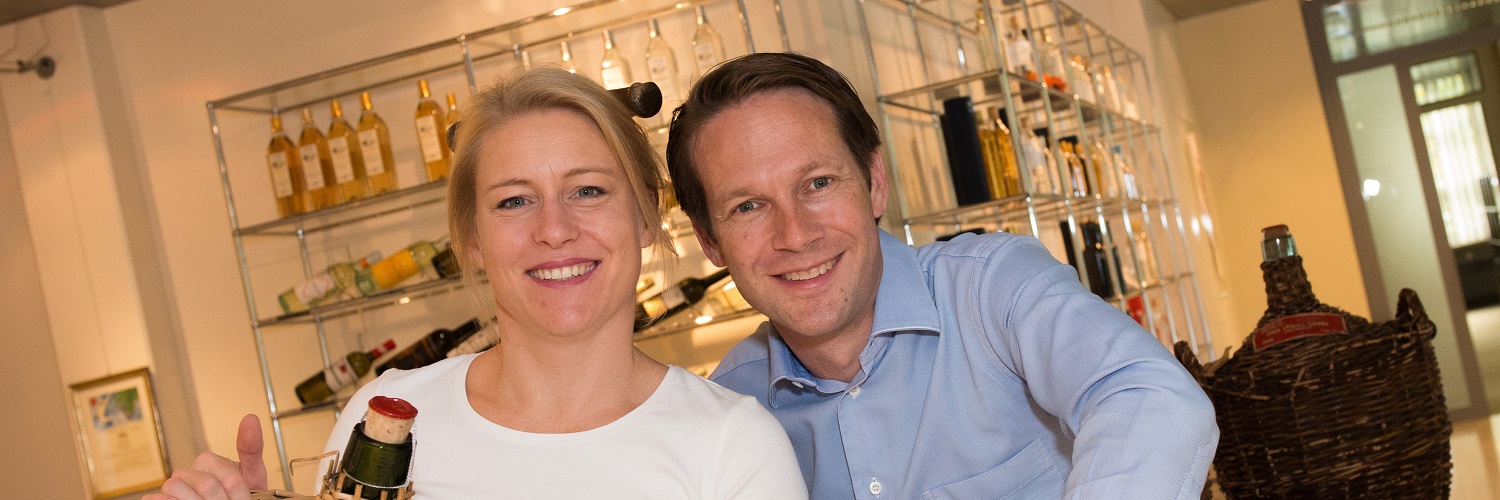 Zwei lächelnde Personen stehen nebeneinander, eine von ihnen hält eine Flasche mit Korken in der Hand. Im Hintergrund sind Regale mit verschiedenen Flaschen und Produkten sichtbar.