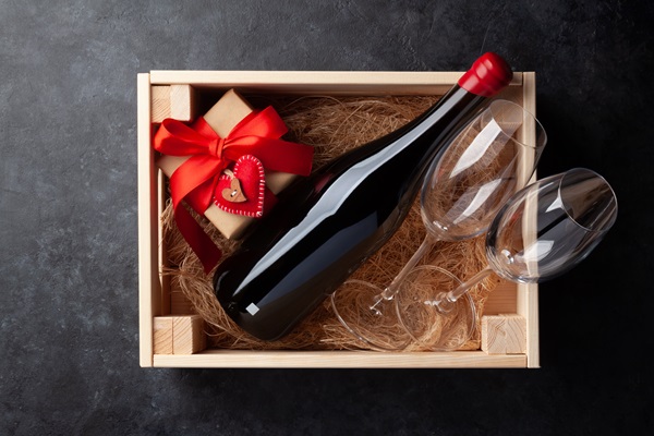 Ein Holzkistchen enthält eine Flasche Rotwein, zwei Weingläser und ein Geschenk mit rotem Band. Das Geschenkpäckchen hat ein Herzornament und ist auf Strohhubbeln platziert.