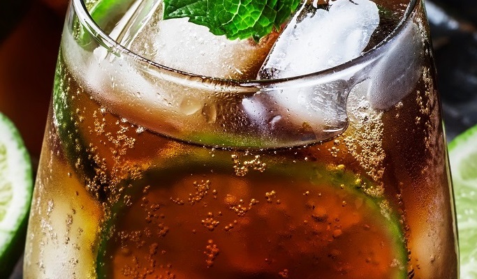 Ein Glas mit einem kohlensäurehaltigen Getränk, gefüllt mit Eiswürfeln und einem Limettenscheibchen. Frische Minzblätter dekorieren das Getränk, das auf einer dunklen Unterlage steht.