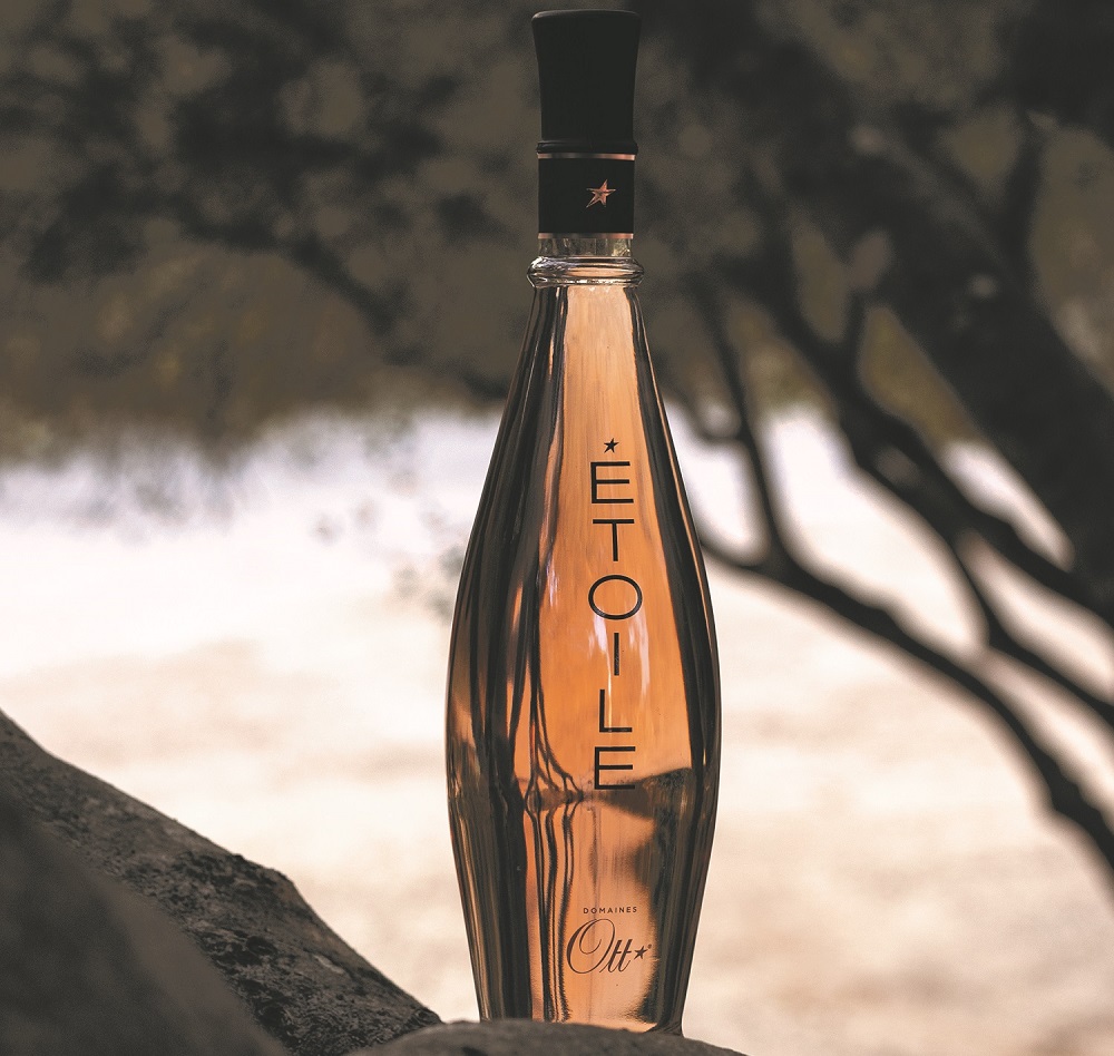 Eine Flasche mit dem Namen -ÉTOILE- steht auf einem Stein, umgeben von unscharfen Bäumen im Hintergrund. Die Flasche hat eine elegante, geschwungene Form und transparenten Roséwein.