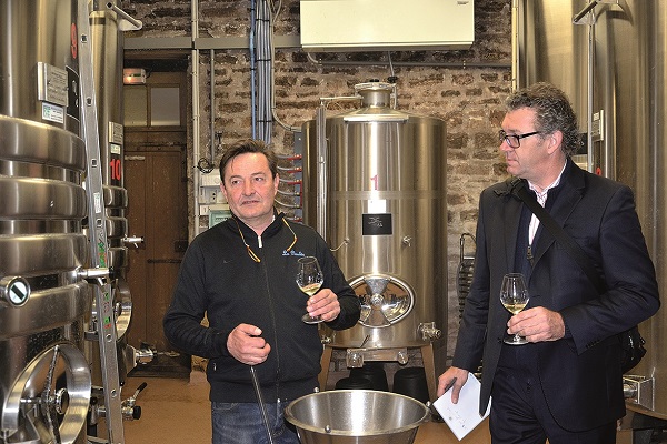 Zwei Männer stehen in einem Weinkeller. Einer hält ein Glas Wein und spricht, während der andere aufmerksam zuhört. Im Hintergrund sind große Metallbehälter und eine Steinmauer sichtbar.