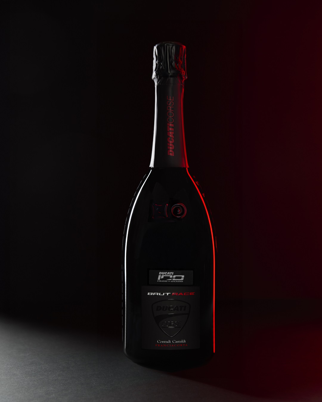 Ein glänzender, schwarzer Champagnerflasche steht vor einem dunklen Hintergrund. Die Flasche hat ein rotes Licht, das sie teilweise umrahmt, und zeigt das Logo von Ducati sowie andere Bieter.