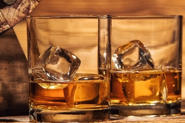 Zwei Gläser mit Whisky, in denen sich jeweils ein Eiswürfel befindet. Sie stehen auf einem rustikalen Holzuntergrund, umgeben von einer warmen, Holztextur.