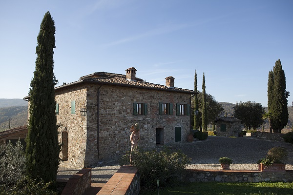 Ein steinernes, zweistöckiges Haus mit grünen Fensterläden steht in einer ländlichen Umgebung. Eine Person steht auf dem Kiesweg davor, umgeben von Zypressen und einem hügeligen Landschaftshorizont.