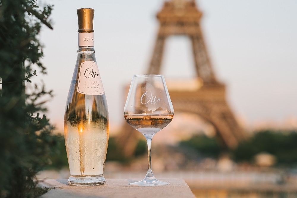 Eine Flasche Roséwein und ein Weinglas stehen auf einem Tisch. Im Hintergrund ist der Eiffelturm, umgeben von einer sanften Abendstimmung und grünem Laub.