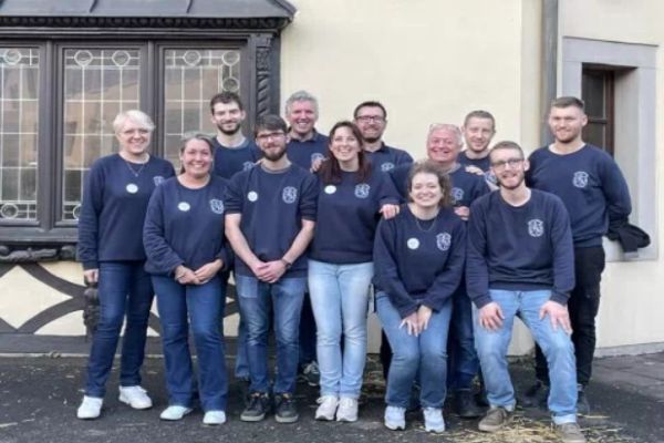 Eine Gruppe von zwölf Personen steht vor einem Gebäude. Alle tragen dunkelblaue Sweatshirts. Einige haben Leggings oder Jeans an. Alle lächeln in die Kamera.