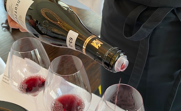 Ein Weinflasche wird gekippt, um roten Wein in ein Glas zu gießen. Die Umgebung zeigt mehrere Gläser mit bereits eingeschenktem Wein auf einem Tisch.