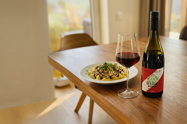 Ein Glaskelch mit Rotwein steht neben einem Teller Pasta, der mit Kräutern garniert ist, auf einem Holztisch in einem hellen Raum.