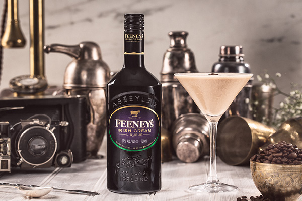 Eine Flasche Feeney-s Irish Cream steht im Vordergrund, neben einem Cocktailglas mit einer milchigen Flüssigkeit. Im Hintergrund befinden sich verschiedene alte Gegenstände und Kaffeebohnen in einer Schale.