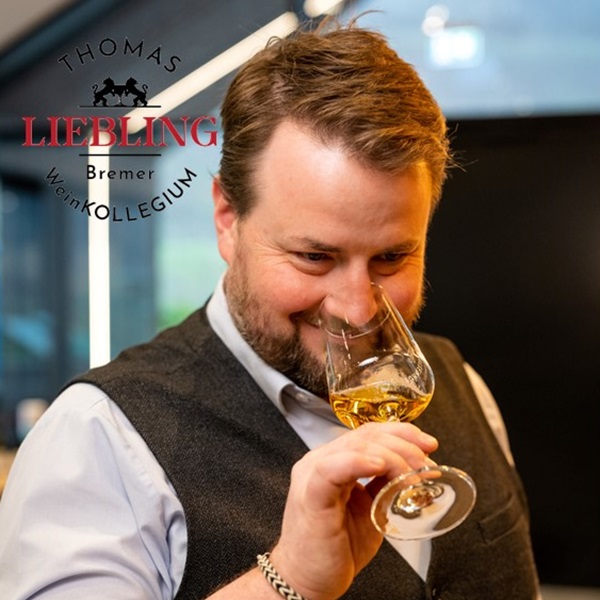 Ein Mann mit einem kurzen Bart und einem grauen Weste hält ein Weinglas, während er den Inhalt daran riecht. Im Hintergrund sind verschwommene Fenster zu sehen.