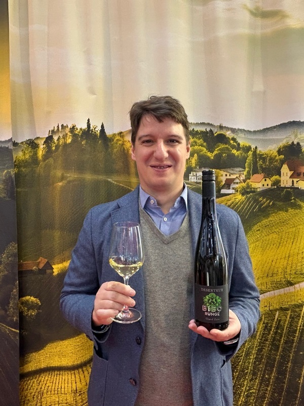 Eine Person hält ein Weinglas mit Weißwein in der linken Hand und eine Flasche Wein in der rechten Hand. Im Hintergrund sind Weinberge und Hügel sichtbar.