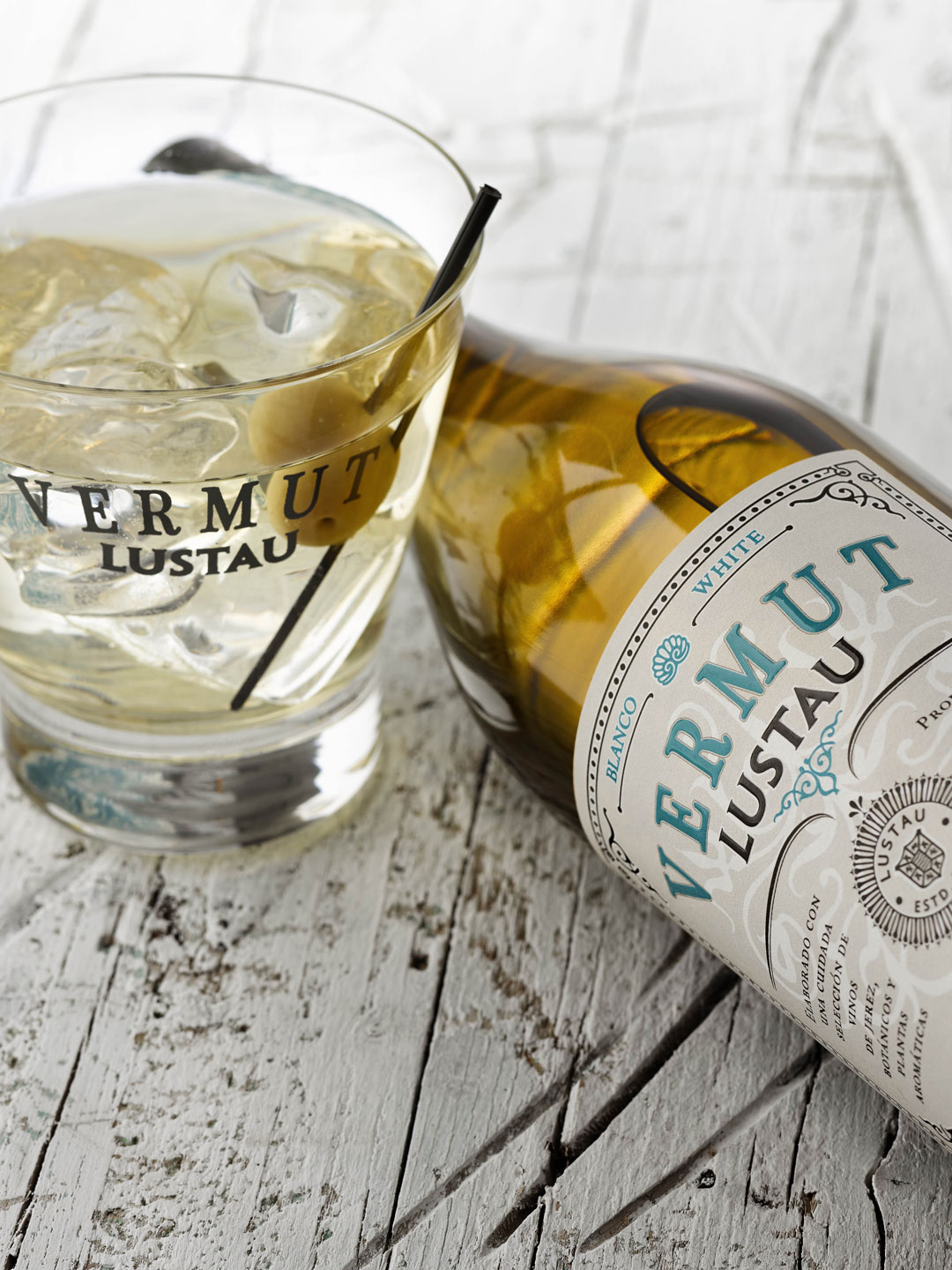 Ein Glas mit Eis und Vermut Lustau sowie einer Olive steht auf einer rustikalen Holzoberfläche. Eine Flasche Vermut Lustau liegt schräg daneben.