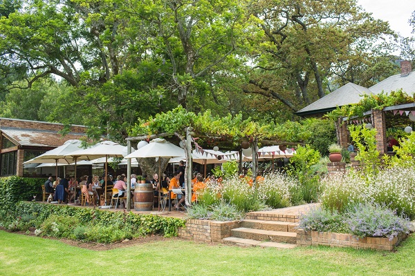 Eine entspannte Restaurantterrasse mit mehreren schattenspendenden Sonnenschirmen. Gäste sitzen an Tischen, umgeben von Bäumen, Blumen und Grünflächen, während das Gebäude im Hintergrund sichtbar ist.