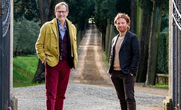 Zwei Männer stehen vor einem geschotterten Weg, umgeben von hohen Zypressen. Der erste trägt eine gelbe Jacke und rote Hosen, der zweite einen schwarzen Blazer.