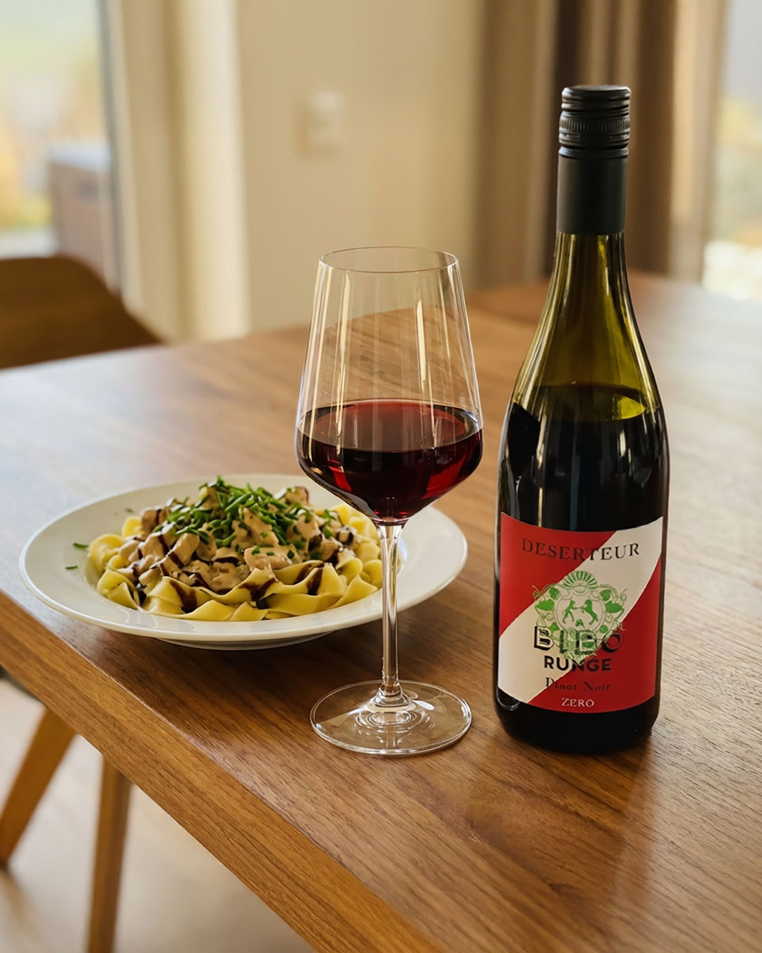 Ein Teller mit Bandnudeln und Hühnchenstücken, garniert mit Schnittlauch, steht auf einem Holztisch. Daneben befindet sich ein Glas Rotwein und eine Flasche mit einem auffälligen Etikett.