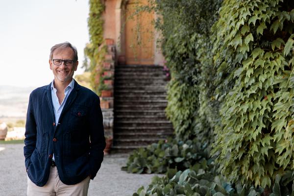 Ein Mann steht lächelnd in einem grünen Garten, umgeben von üppigem Bewuchs und Treppen, die zu einem Gebäude führen. Er trägt einen blauen Blazer und helle Hosen.