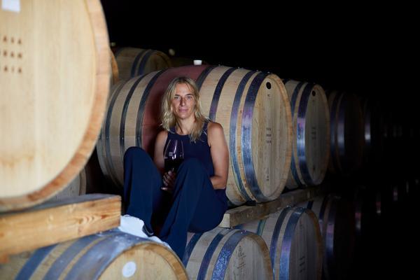 Eine Frau sitzt auf einem Fass in einem Weinkeller, hält ein Weinglas in der Hand. Um sie herum stehen mehrere Holzfässer in einem dunklen, gedämpften Licht.