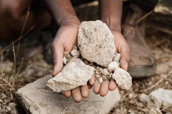 Hände halten mehrere Steine, während sich der Mensch auf einem unebenen Boden befindet. Im Hintergrund sind unscharfe Erd- und Pflanzenreste zu sehen.
