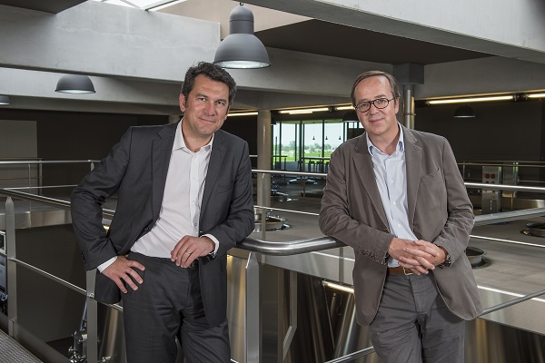 Zwei Männer stehen in einem modernen Raum mit großen Fenstern und industriellem Design. Sie lehnen an einem Geländer und lächeln in die Kamera.