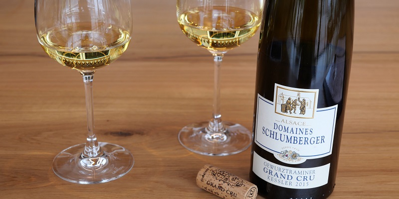 Zwei Weingläser mit hellgelbem Wein stehen auf einem Holztisch neben einer Flasche Gewürztraminer Grand Cru Kessler 2015 und einem Korken.
