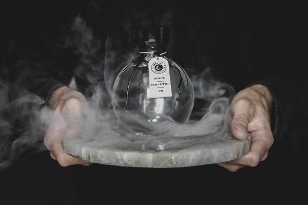 Eine Person hält eine runde, transparente Flasche mit einem Etikett, die auf einem grauen, runden Steinuntersetzer steht. Zarte Rauchschwaden umgeben die Flasche in einer dunklen Umgebung.