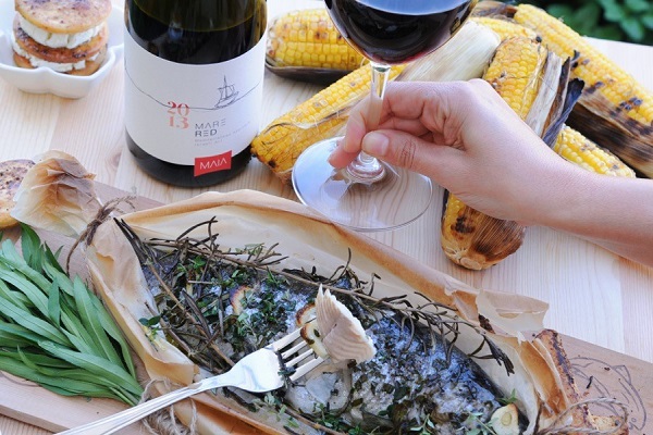 Ein handgehaltenes Weinglas mit rotem Wein wird neben einem Teller mit Fisch, der mit Kräutern garniert ist, präsentiert. Im Hintergrund befindet sich gekochter Mais.