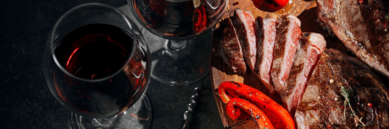 Zwei Hände mit Weingläsern stoßen an, während im Hintergrund Teller mit Salat, Fleisch und einer Birne zu sehen sind. Die Tischoberfläche ist aus Holz und dekoriert mit frischen Blumen.