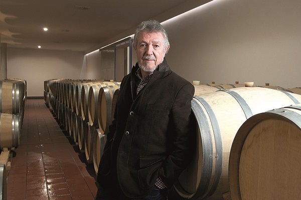 Ein Mann steht in einem Weinkeller, umgeben von Holzfässern. Er trägt einen dunklen Mantel und hat graues Haar. Der Raum ist beleuchtet und hat einen modernen, sauberen Look.