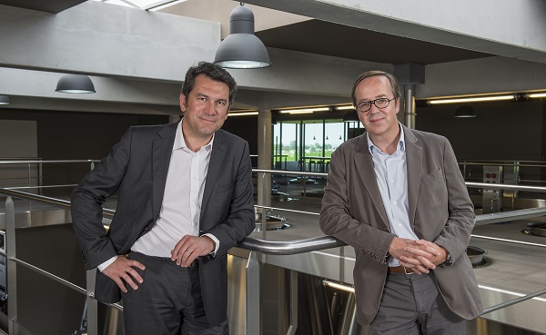 Zwei Männer stehen in einem modernen Innenraum, der von natürlichem Licht durch große Fenster erleuchtet wird. Sie lehnen lässig an einem Geländer und blicken freundlich in die Kamera.