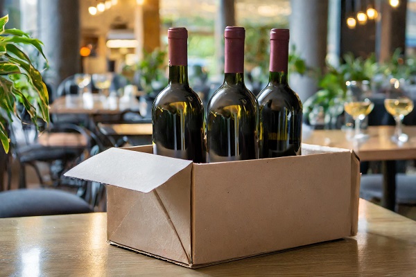 Drei Flaschen Wein stehen in einem geöffneten Karton auf einem Tisch in einem Restaurant. Der Hintergrund zeigt eine einladende Atmosphäre mit Pflanzen und gedeckten Tischen.