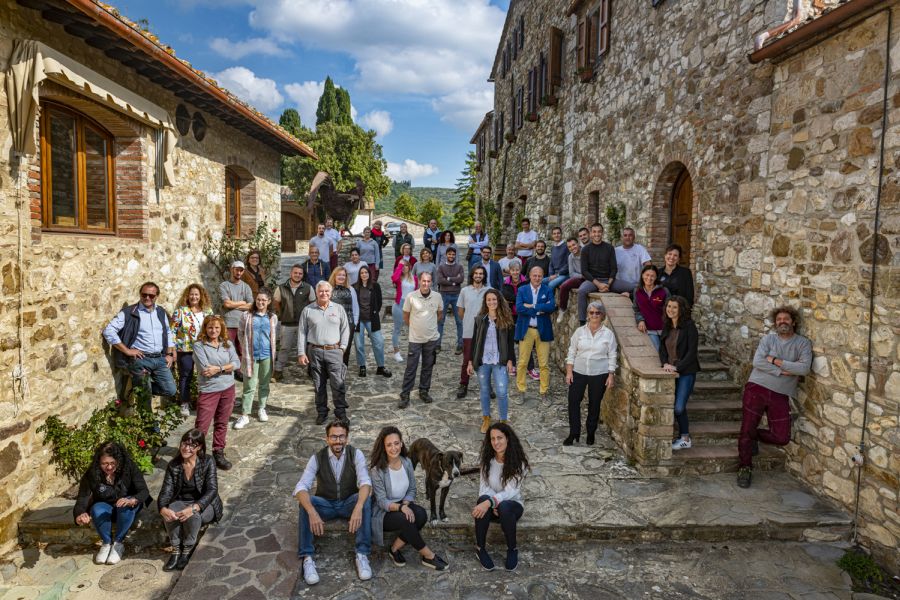 Eine Gruppe von etwa 50 Personen steht auf einem gepflasterten Weg in einer malerischen, von Steinmauern umgebenen Umgebung. Die Szenerie zeigt einige Bäume und Gebäude im Hintergrund unter einem klaren Himmel.