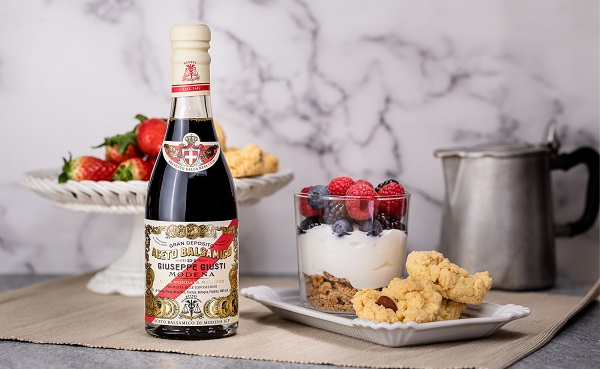Eine Flasche Balsamico-Essig steht neben einem Glas mit Joghurt, Beeren und süßem Gebäck auf einem Tisch. Im Hintergrund befindet sich eine Schale mit frischen Erdbeeren.