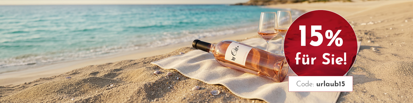 Eine Flasche Roséwein steht neben zwei Weingläsern auf einem weißen Tuch am Sandstrand. Im Hintergrund sind das Meer und der klare Himmel sichtbar. Text: -15% für Sie! Code: urllaub15-.
