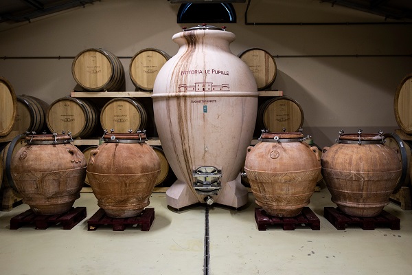 Große Lektionen aus Ton stehen in einer Weinkelterei, umgeben von Holzfässern, auf dem Boden. Der Raum hat eine funktionale, industriell anmutende Atmosphäre.