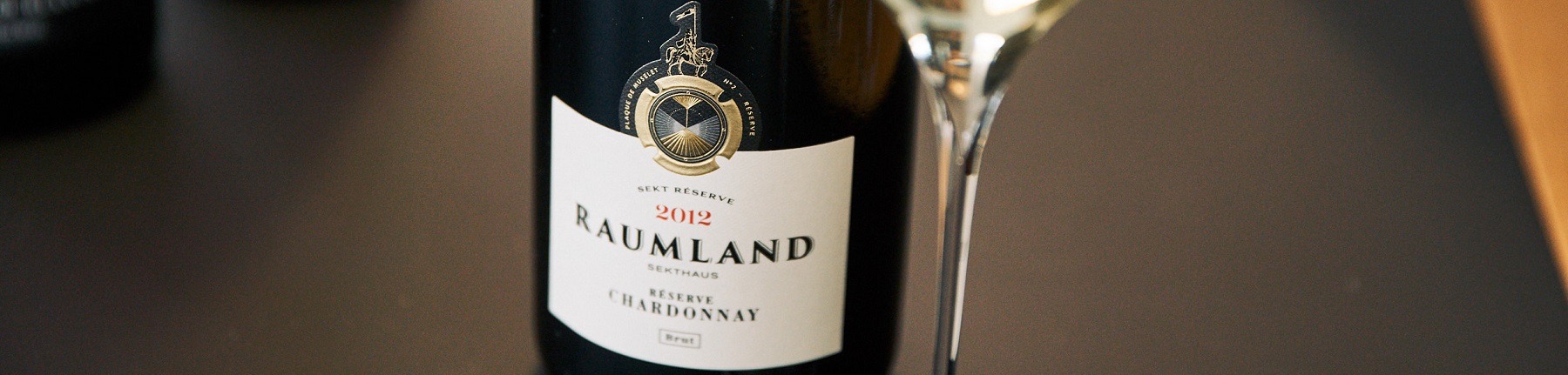 Raumland Eine Flasche Sekt mit dem Etikett -Raumland 2012 Reserve Chardonnay- steht auf einem Tisch. Daneben befindet sich ein Glas, das möglicherweise für den Sekt gedacht ist.